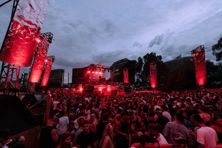Large crowd dancing at Sidebyside Festival under red stage lighting at dusk with city skyline in background.